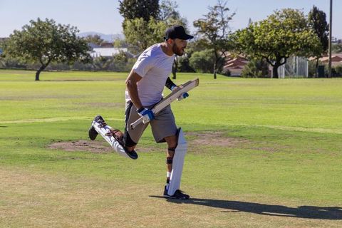 Male cricketer sprinting between wickets carrying bat wearing pads and gloves on sunny field