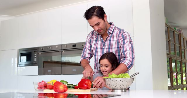 Father slicing vegetables while daughter watches in modern kitchen. Useful for content about family bonds, cooking together, healthy living, parental engagement, and home meals. Ideal for blogs, advertisements, and educational materials related to family and nutrition.