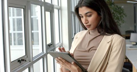 Indian businesswoman standing by window using tablet in modern minimalist office daylight