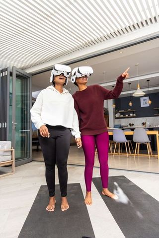 Black women holding hands wearing vr headsets on patio yoga mats practicing mindful wellness