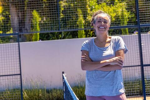 Smiling middle-aged african american woman standing by net on sunlit court, casual athletic