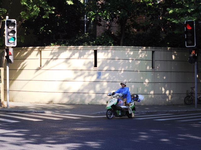 Picture showing a person riding a green scooter, capturing everyday urban commuting at an intersection marked with traffic lights indicating the stoplight. The background features a shadowed wall and trees, giving the scene a warm evening vibe. Perfect for use in campaigns focusing on city commuting, transportation safety, or urban lifestyle.