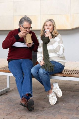 Mature couple sitting on urban bench eating takeout and sipping coffee in city plaza