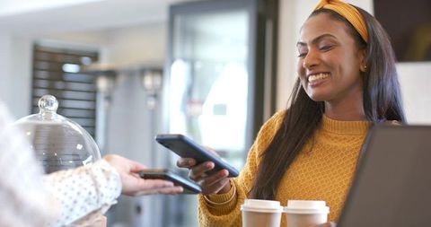 Indian woman making contactless mobile payment at coffee counter in mustard sweater carrying two cup