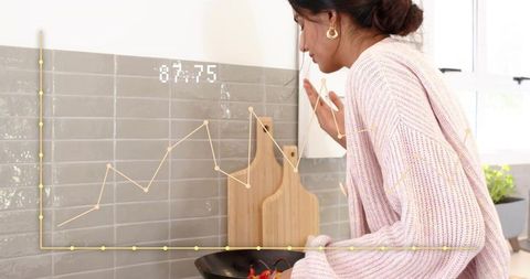 Indian woman in pink knit sweater leaning and checking frying pan with peppers in bright kitchen