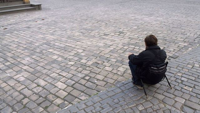 Sitting man on folding stool on expansive cobblestone plaza contemplating urban solitude
