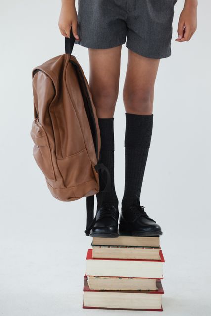 Schoolboy in uniform standing on a stack of books while holding a backpack. This image can be used for educational materials, back-to-school promotions, academic success themes, and childhood learning concepts.