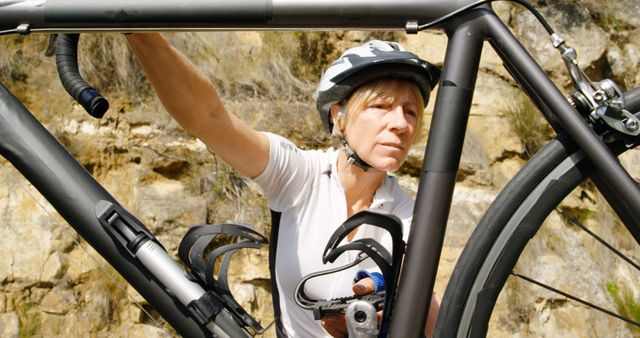 A woman is adjusting a bicycle outdoors on a sunny day, with rocky nature in the background. She is concentrated on fixing her bike, wearing a helmet and cycling gear. This image is suitable for topics related to outdoor activities, cycling, sports maintenance, and active lifestyle promotions.