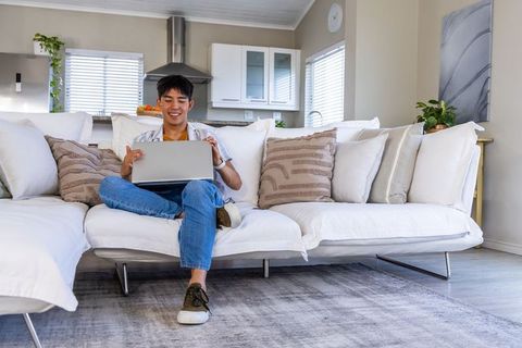 Young asian man working on laptop on white sectional sofa in bright modern living room