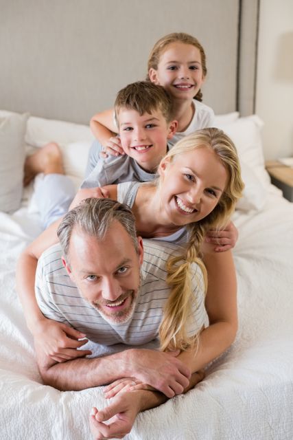 This image shows a joyful family of four, with parents and children stacking on top of each other on a bed. They are smiling and appear to be enjoying a playful moment together. This photo can be used for family-oriented content, advertisements, or articles about family bonding, home life, and happiness.