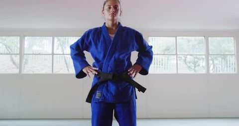 Confident young female martial artist standing in blue gi with black belt in bright dojo