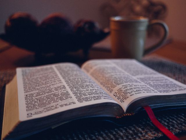 Image displays an open Bible with a red bookmark, placed on a table. A blurred mug in the background suggests a cozy, warm setting ideal for reading and studying. Ideal for use in religious content, articles on spirituality, blog posts on faith, or advertisements for faith-based products. The warm tones and soft lighting contribute to a peaceful atmosphere.