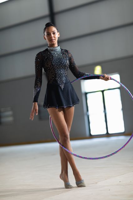 Biracial female gymnast practicing with a hula hoop in a gym, looking into the camera. She is training hard for an upcoming competition. This image can be used for articles on gymnastics, sports training, athleticism, and determination. It is also suitable for promoting gymnastics events, sportswear, and fitness programs.