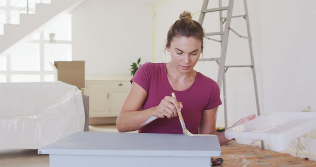A woman is painting a piece of furniture with a small brush in a bright and airy living room. There is a ladder in the background and some cardboard boxes, suggesting a home renovation project. This image could be used for articles or advertisements about home improvement, DIY projects, interior design, or lifestyle blogs focused on personal craftsmanship and creativity.