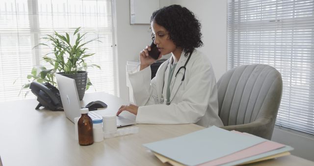 Doctor in white coat with stethoscope talking on phone while using laptop at desk. Surroundings include patient files, medicine bottles and plant. Ideal for use in healthcare promotions, medical blogs, office productivity content, or doctor-patient communication visuals.