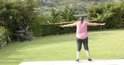 Mature woman stretching arms on patio edge facing manicured backyard lawn, home fitness