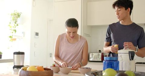 Sunlit modern kitchen couple preparing fresh fruit and bowls, slicing strawberries together