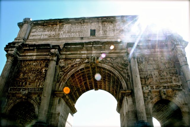 This image showcases an ancient Roman archway with engravings at the top, basking in bright sunlight that creates light flare effects. Such visuals are ideal for illustrating historical articles, travel blogs, educational content about ancient Rome, and marketing materials for tourism agencies.
