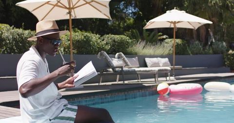 Man in straw hat reading and drinking poolside with inflatable floats and umbrellas
