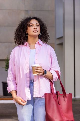 Confident woman standing with coffee and red tote wearing pink shirt and smartwatch