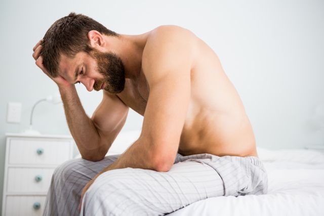 This image depicts a shirtless man sitting on a bed with his hand on his head, appearing to be in deep thought or distress. It can be used in articles or campaigns related to mental health, depression, anxiety, personal struggles, and emotional well-being. It is also suitable for illustrating themes of solitude, contemplation, and stress.
