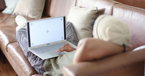 African American man reclining on leather sofa using laptop for remote work and relaxation
