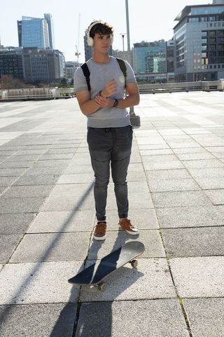 Teen male adjusting wristwatch beside skateboard in sunny urban plaza with headphones