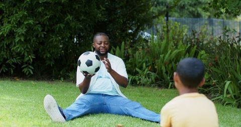 African american father passing soccer ball to son sitting on green backyard lawn