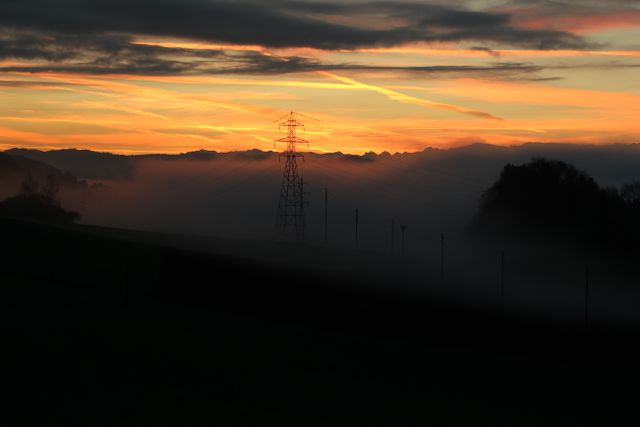 This stunning scene captures an electricity pylon silhouette against a vivid sunset sky, with mist creating a dramatic horizon line. Ideal for publications, presentations, or backgrounds that require tranquil yet powerful visual impact, illustrating energy, infrastructure, and natural beauty.