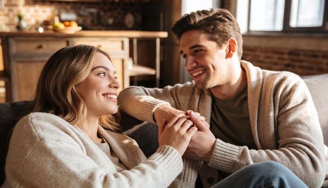 Couple holding hands on couch in cozy kitchen wearing knit cardigans, smiling warmly