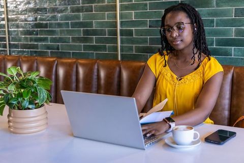 African american woman working on laptop reviewing notes in modern cafe with coffee