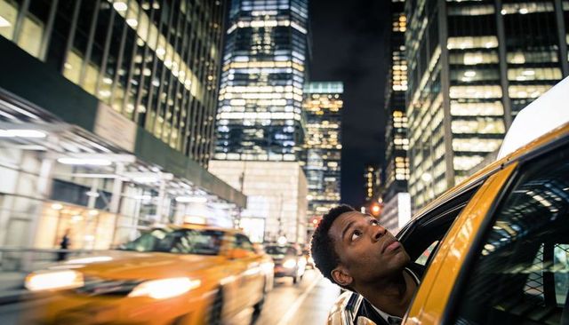 Man leaning out of yellow taxi at night in bustling downtown, motion blur city lights