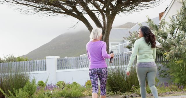Two women are walking together outdoors wearing activewear. One has white hair and the other has dark long hair. They are carrying water bottles. There are houses and a mountain in the background. Perfect for illustrating healthy lifestyle, fitness, and outdoor activities.