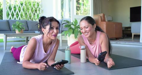 African american and indian women sharing laughter on yoga mats using smartphones