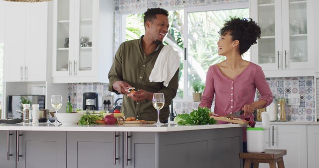 This image captures an African American couple enjoying each other's company while cooking and sipping wine at home. The mood is cheerful and relaxed, presenting a warm and loving domestic atmosphere. This can be used in marketing materials for dating services, cooking classes, food and wine brands, home living advertisements, or lifestyle blogs. Its theme of connection and togetherness makes it suitable for illustrating romantic or family-oriented campaigns.