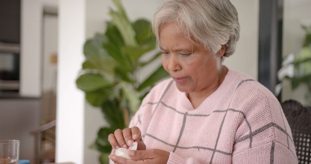 Senior woman taking medication at home. She appears focused while holding a container of pills in her hands. Suitable for health care, elderly care, medical treatment, and wellness-related concepts.