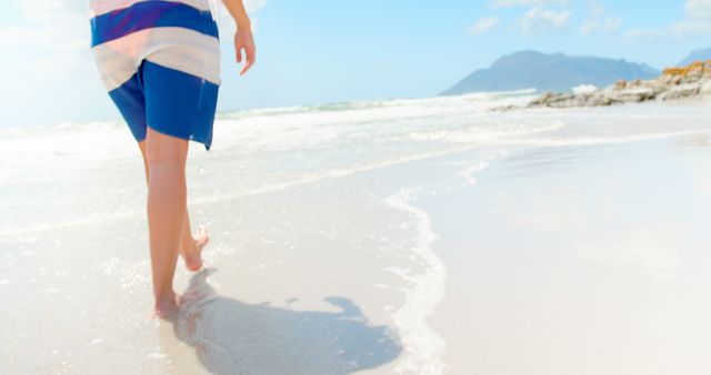 Person walking barefoot along a sunny beach, wearing blue shorts. Ocean waves gently hitting the shore, creating a serene and tranquil environment. Ideal for use in travel advertisements, relaxation and wellness content, and summertime promotions.