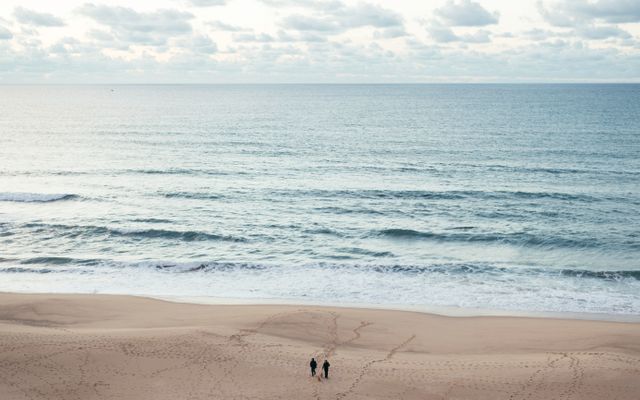 Two people walking along a serene beach with an expansive ocean view and cloud-studded sky. Ideal for promoting travel destinations, vacation spots, nature experiences, and relaxation. Use this for travel blogs, tourism ads, and wellness articles.