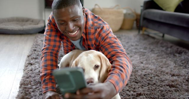 A man taking a selfie with his dog while lying on a carpeted floor in a modern living room. Both are relaxed and happy, showcasing a strong friendship and bond. This image can be used for articles, blogs, and advertisements about pet ownership, bonding moments, modern tech usage, and lifestyle.
