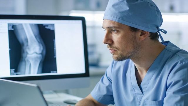 Radiologist reviewing knee x-ray on monitor wearing scrubs and surgical cap