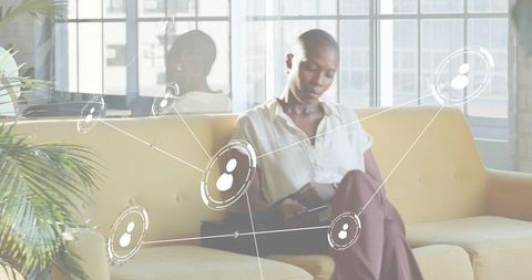 Professional relaxing on mustard sofa using tablet with network overlay in sunlit lounge