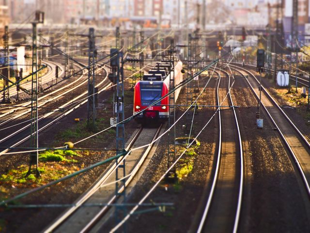 This image depicts a red commuter train gliding on a set of railway tracks in an urban environment. Ideal for use in transportation-related projects, urban development presentations, travel brochures, and infrastructure planning documents. Perfect for illustrating concepts related to city connectivity and modern transit solutions.