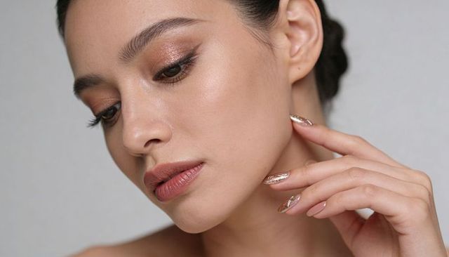 Bronze glam closeup asian woman touching jawline with metallic foil nail art and bare shoulders