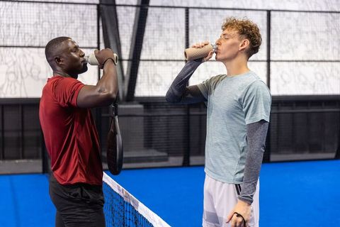 Diverse male partners drinking from bottles on indoor padel court after friendly match