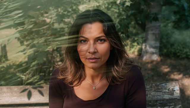 Woman sitting on wooden bench in dappled sunlight wearing pendant necklace and gazing