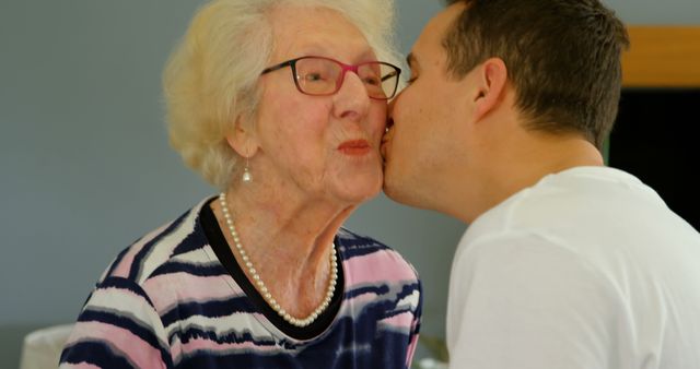 Close up of an adult son kissing his retired elder mother in living room at home. The father is holding the hands of the grandmother.
