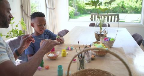 Father and son painting easter eggs at dining table during sunny family craft bonding