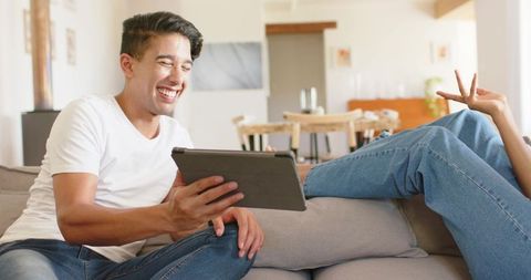 Laughing couple relaxing on sofa using tablet while partner flashes peace sign