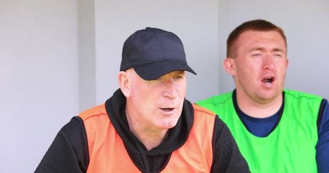 Male team players sitting in dugout calling to field wearing training bibs and cap