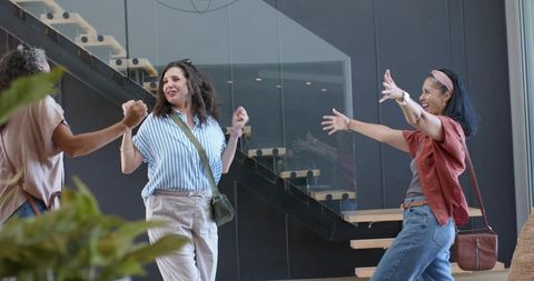 Women reuniting in modern lobby embracing and laughing by glass staircase